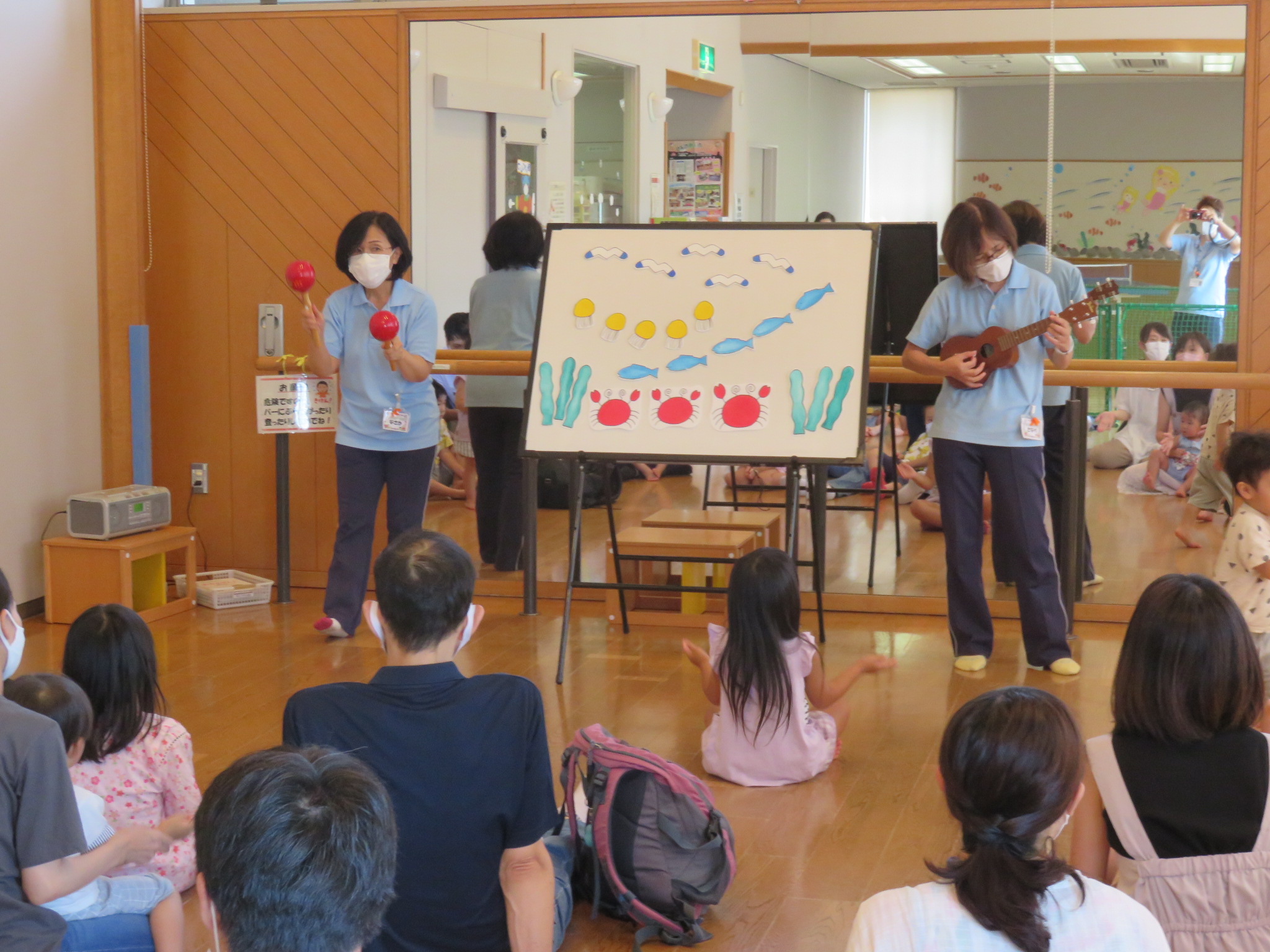 児童館職員が親子連れの前でマラカスやギターを使って歌ったりお話ししたりしている写真